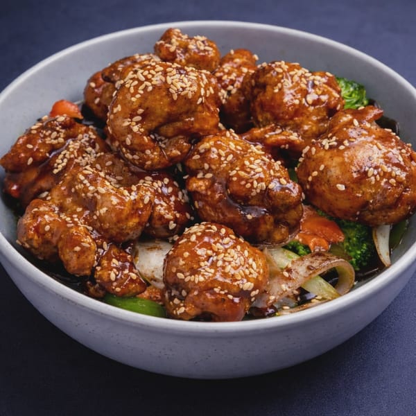 Sesame chicken with broccoli, onions, and carrots in a light gray bowl on a dark background.