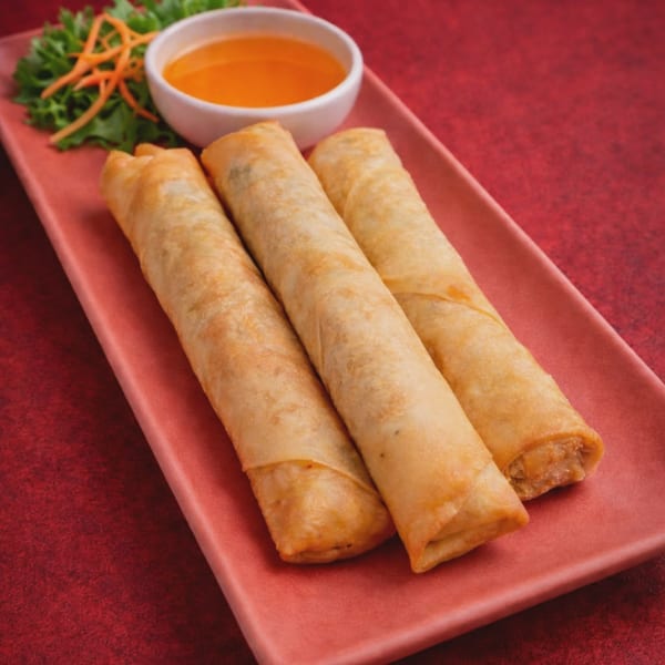 Three golden spring rolls on a pink plate with a side of orange dipping sauce and garnish.
